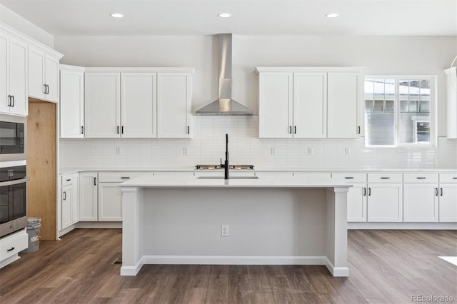 kitchen with appliances with stainless steel finishes, white cabinetry, hardwood / wood-style flooring, a kitchen island with sink, and wall chimney exhaust hood