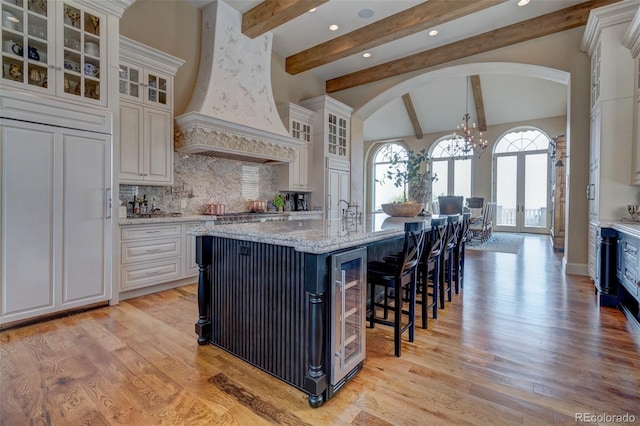 kitchen with premium range hood, a center island with sink, backsplash, light stone countertops, and light hardwood / wood-style flooring