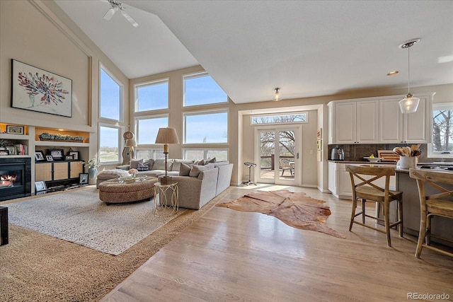 living area featuring a glass covered fireplace, light wood-type flooring, a towering ceiling, and ceiling fan