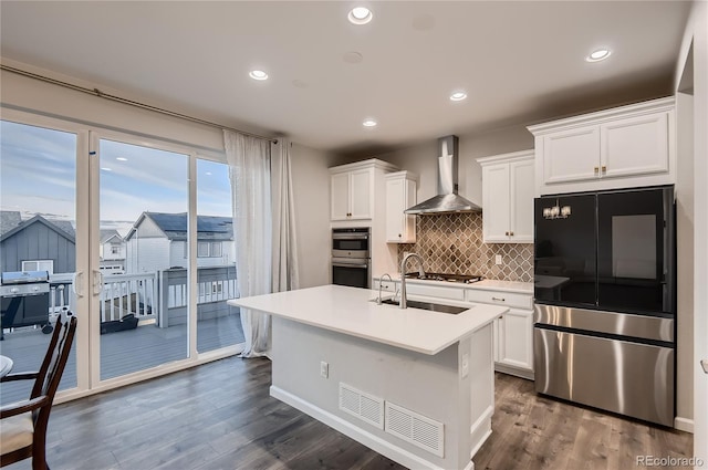 kitchen featuring wall chimney range hood, sink, white cabinetry, stainless steel appliances, and a center island with sink