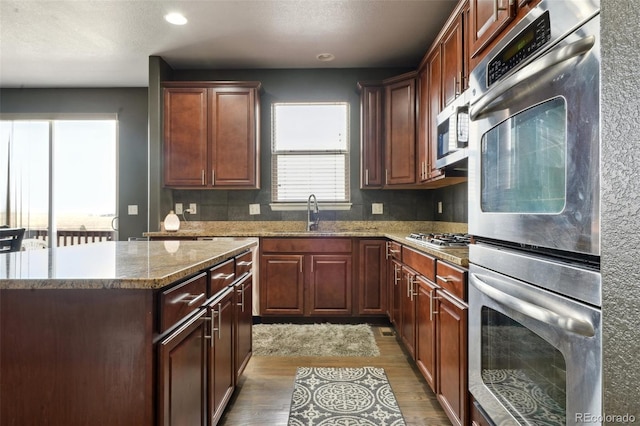 kitchen with wood finished floors, appliances with stainless steel finishes, a sink, a kitchen island, and light stone countertops