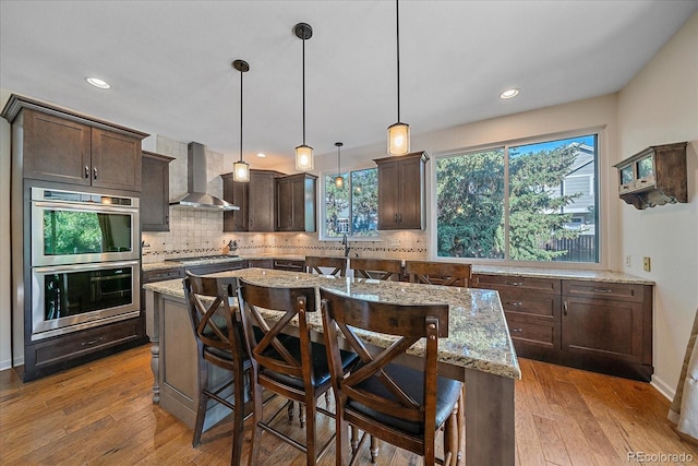 10421 Kennemere Ln, Parker, CO 80134 - kitchen featuring wall chimney exhaust hood, stainless steel double oven, dark brown cabinetry, a center island, and light stone countertops
