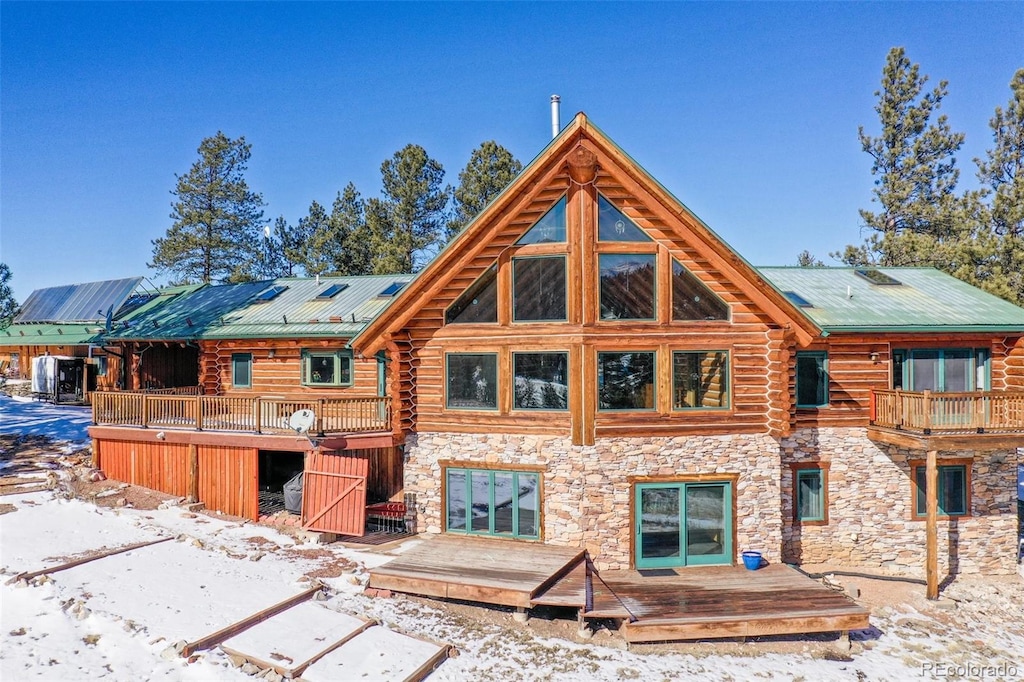 snow covered back of property featuring stone siding, metal roof, log exterior, and a wooden deck