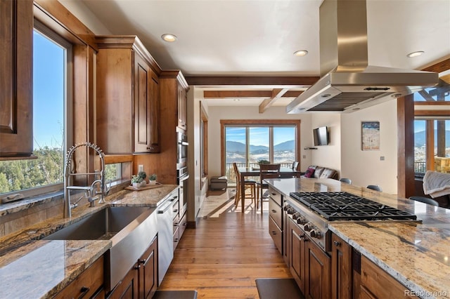 kitchen featuring light wood-type flooring, light stone counters, stainless steel appliances, island range hood, and beam ceiling