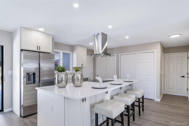 kitchen with a kitchen island with sink, a kitchen breakfast bar, stainless steel refrigerator with ice dispenser, island range hood, and white cabinets