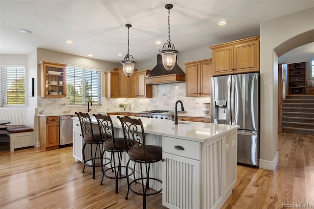 kitchen featuring appliances with stainless steel finishes, light hardwood / wood-style floors, custom exhaust hood, decorative light fixtures, and a kitchen island with sink