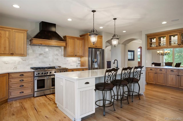 kitchen featuring custom exhaust hood, an island with sink, appliances with stainless steel finishes, a kitchen breakfast bar, and light hardwood / wood-style floors