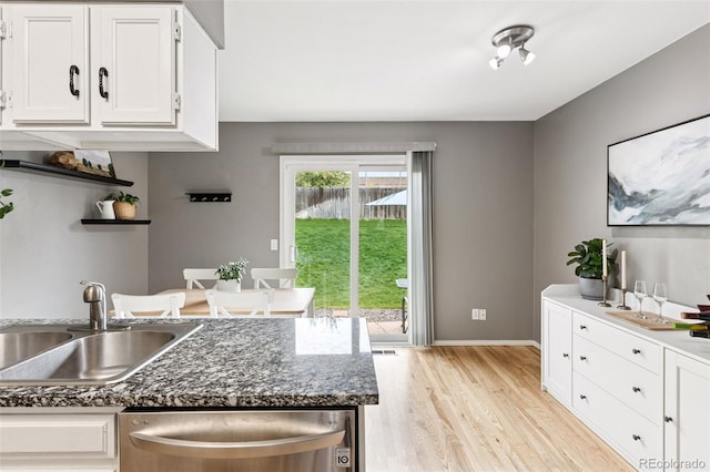 kitchen with stainless steel dishwasher, a sink, white cabinets, light wood finished floors, and dark stone countertops