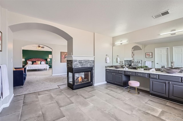 ensuite bathroom featuring double vanity, a sink, visible vents, and a multi sided fireplace