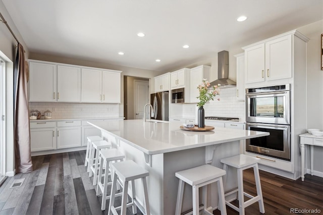 kitchen featuring white cabinetry, light countertops, appliances with stainless steel finishes, wall chimney range hood, and a kitchen bar