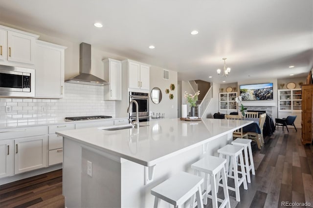 kitchen featuring an island with sink, appliances with stainless steel finishes, wall chimney range hood, a fireplace, and a sink