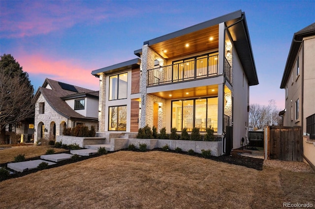 back of house at dusk with a balcony, stone siding, and stucco siding