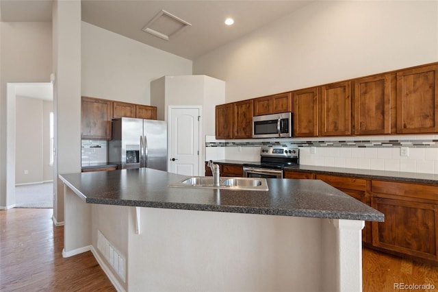 kitchen featuring stainless steel appliances, a towering ceiling, dark countertops, a kitchen island with sink, and recessed lighting