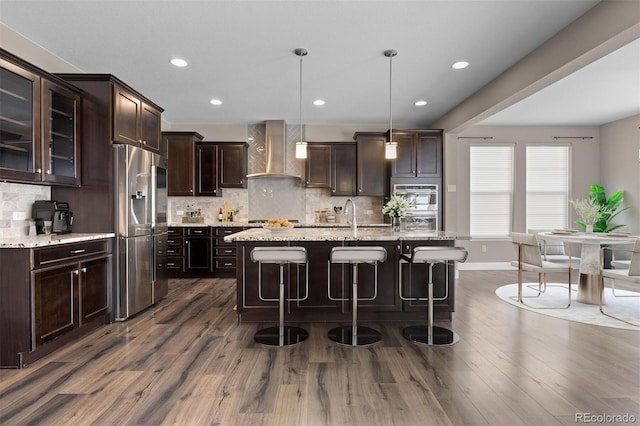 kitchen with pendant lighting, an island with sink, stainless steel appliances, and wall chimney range hood