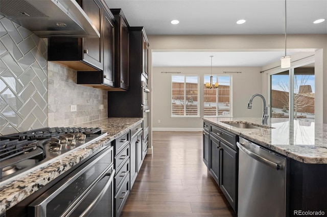 kitchen featuring sink, stainless steel appliances, wall chimney range hood, light stone counters, and decorative light fixtures