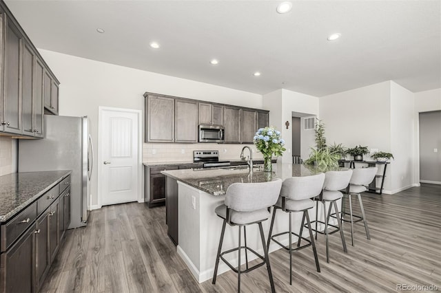 kitchen featuring sink, tasteful backsplash, appliances with stainless steel finishes, an island with sink, and dark stone counters