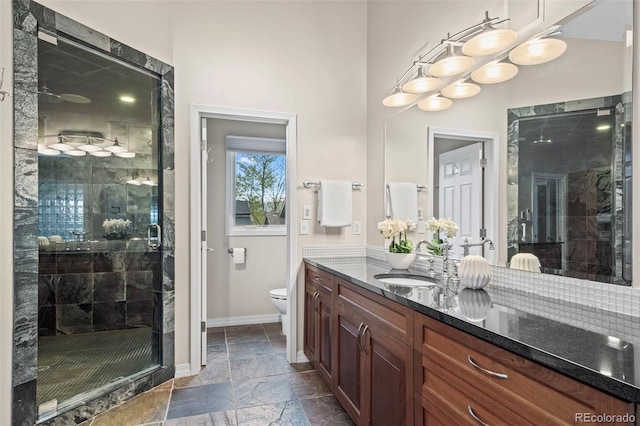 full bath with toilet, vanity, a stall shower, stone tile floors, and baseboards