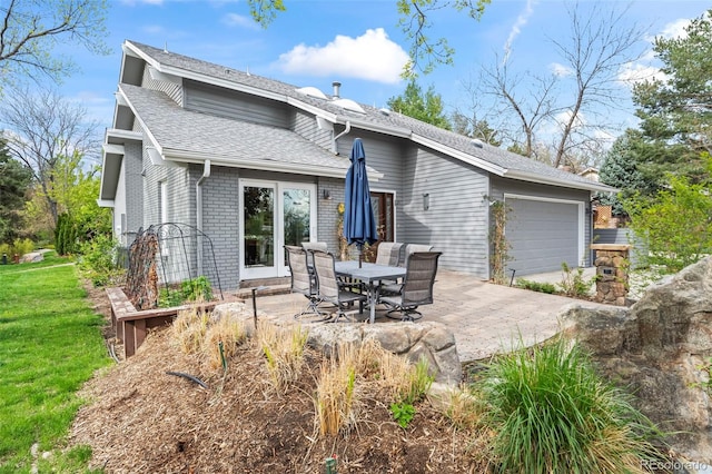 rear view of house with an attached garage, a lawn, a shingled roof, brick siding, and a patio area