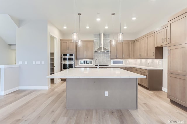 kitchen featuring wall chimney exhaust hood, a center island with sink, pendant lighting, stainless steel double oven, and decorative backsplash