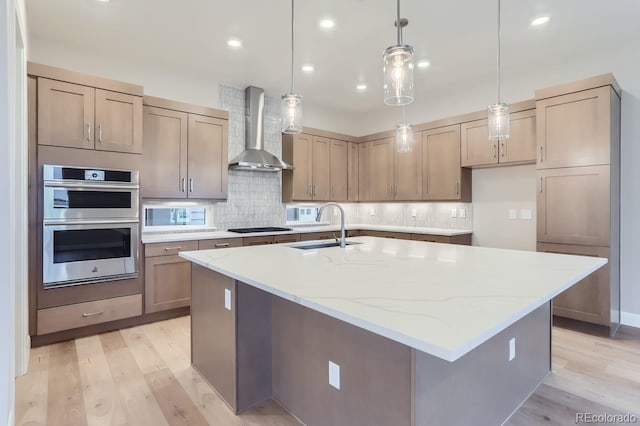 kitchen featuring wall chimney range hood, sink, gas cooktop, light stone counters, and a center island with sink