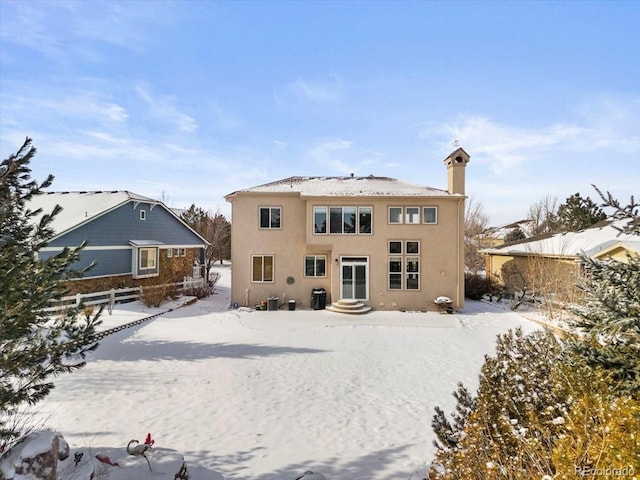 snow covered house with stucco siding, entry steps, a chimney, and fence