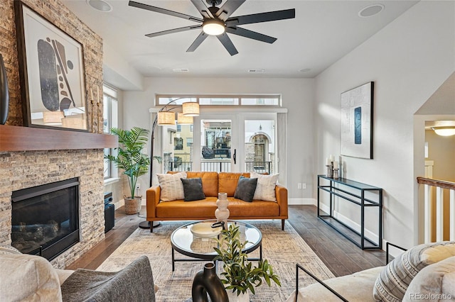 living room with a stone fireplace, ceiling fan, baseboards, and dark wood finished floors