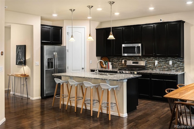 kitchen with pendant lighting, dark hardwood / wood-style floors, an island with sink, backsplash, and appliances with stainless steel finishes