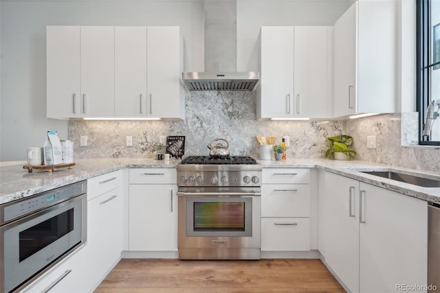 kitchen with wall chimney range hood, light stone countertops, white cabinetry, light hardwood / wood-style flooring, and stainless steel appliances