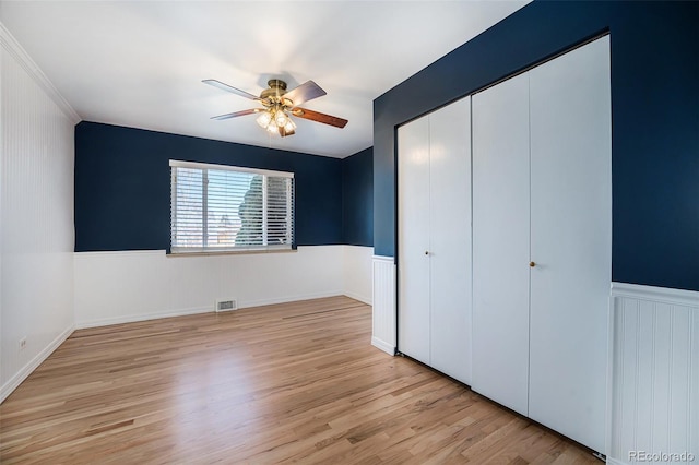 unfurnished bedroom featuring visible vents, a ceiling fan, a wainscoted wall, light wood-style flooring, and a closet