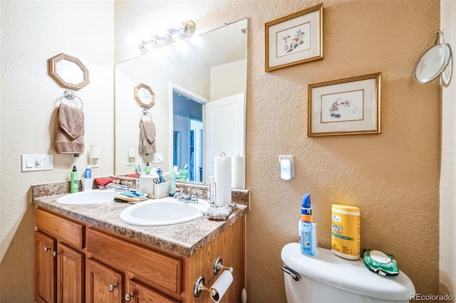 half bath featuring a textured wall, double vanity, a sink, and toilet