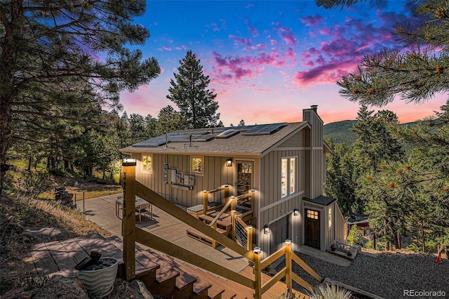 32783 Lodgepole Cir, Evergreen, CO 80439 - back of property at dusk featuring roof mounted solar panels, a chimney, a deck, board and batten siding, and roof with shingles