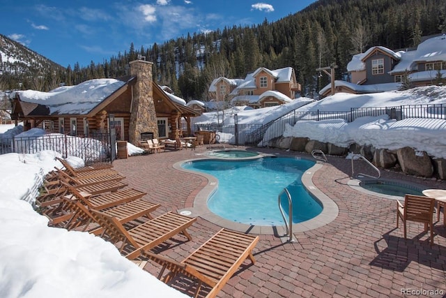 snow covered pool featuring a fenced in pool, a fenced backyard, a view of trees, and an in ground hot tub