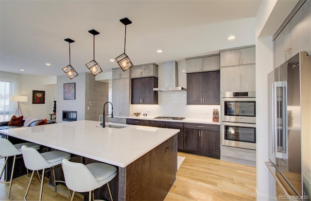 kitchen with sink, wall chimney exhaust hood, an island with sink, appliances with stainless steel finishes, and decorative light fixtures