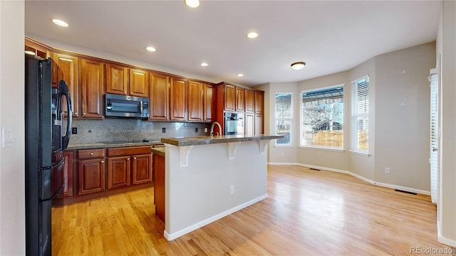 kitchen featuring light hardwood / wood-style floors, a kitchen bar, a center island with sink, tasteful backsplash, and black appliances