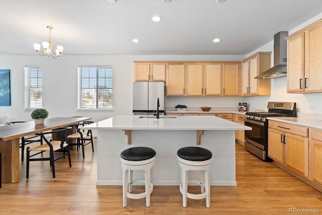 kitchen featuring sink, decorative light fixtures, appliances with stainless steel finishes, a kitchen island with sink, and wall chimney range hood