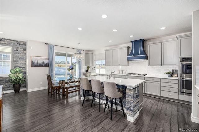 kitchen with gray cabinetry, stainless steel appliances, wall chimney exhaust hood, and an island with sink