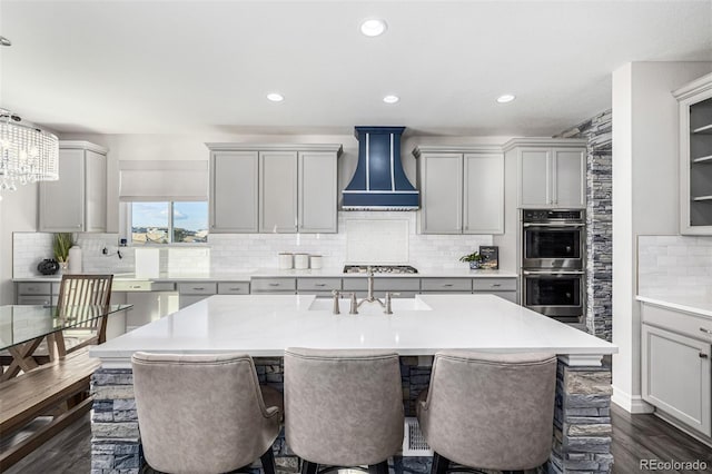 kitchen with appliances with stainless steel finishes, gray cabinetry, an island with sink, and wall chimney range hood