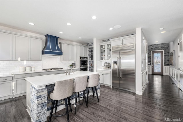 kitchen featuring wall chimney range hood, dark wood-type flooring, appliances with stainless steel finishes, a kitchen breakfast bar, and an island with sink