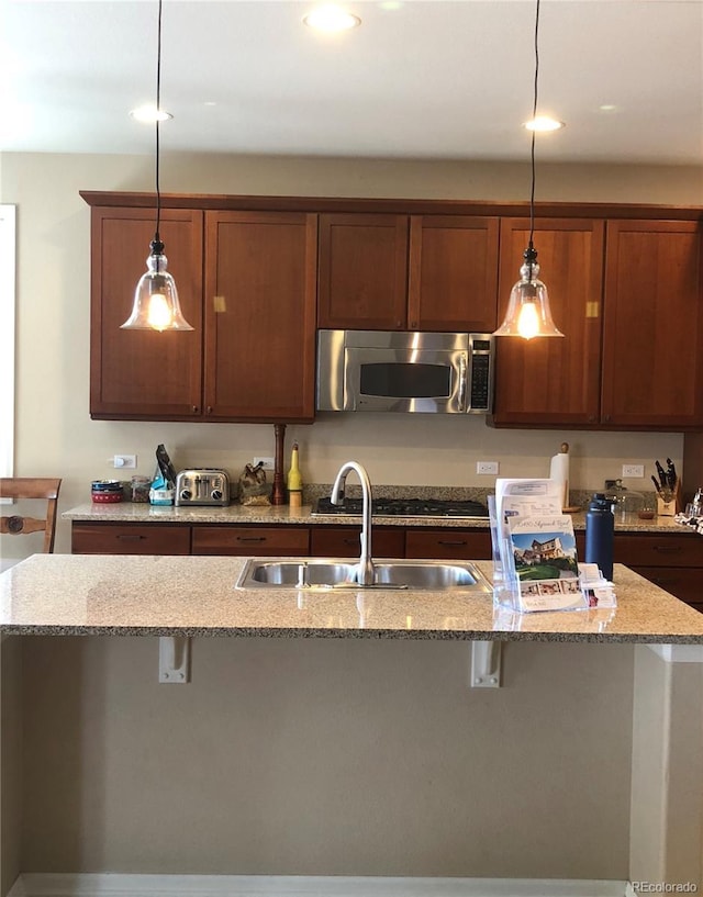 kitchen with decorative light fixtures, a sink, light stone counters, stainless steel microwave, and a breakfast bar