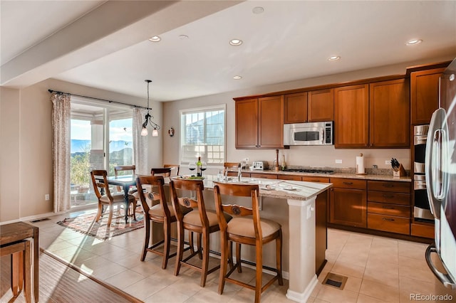 kitchen with brown cabinets, light tile patterned flooring, light stone countertops, an island with sink, and stainless steel appliances