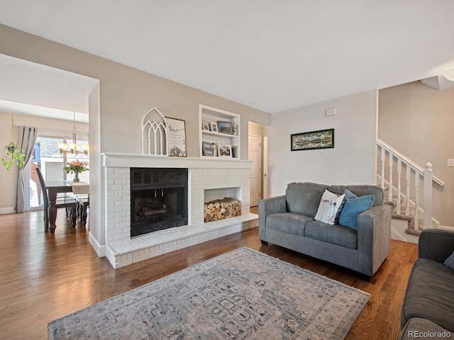 living area featuring built in shelves, a notable chandelier, wood finished floors, stairway, and a brick fireplace