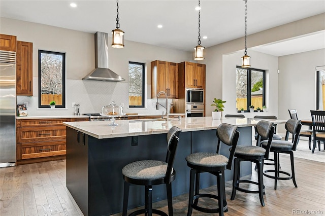 kitchen featuring an island with sink, a sink, brown cabinetry, wall chimney range hood, and built in appliances