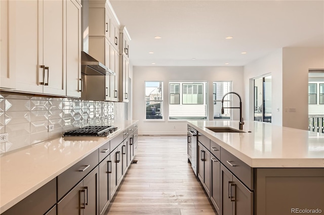 kitchen with sink, wall chimney range hood, light hardwood / wood-style floors, stainless steel gas stovetop, and a center island with sink