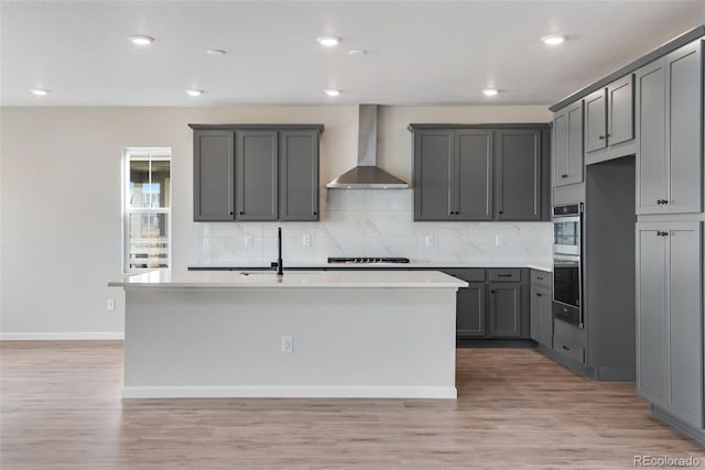 kitchen featuring a center island with sink, wall chimney exhaust hood, a sink, gray cabinets, and backsplash