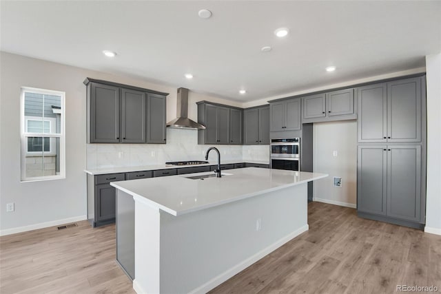 kitchen featuring light wood-style flooring, a sink, visible vents, wall chimney range hood, and a center island with sink