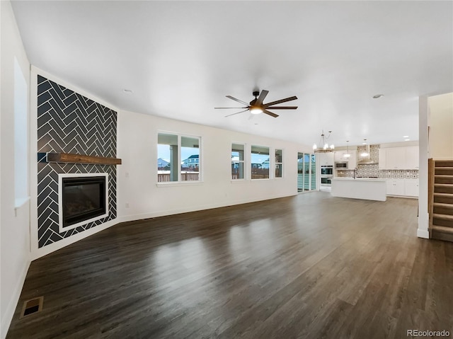 unfurnished living room featuring ceiling fan with notable chandelier, a fireplace, visible vents, baseboards, and dark wood-style floors