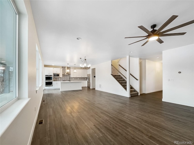 unfurnished living room featuring baseboards, visible vents, dark wood finished floors, stairway, and ceiling fan with notable chandelier