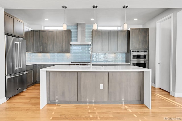 kitchen with appliances with stainless steel finishes, an island with sink, decorative light fixtures, and wall chimney range hood