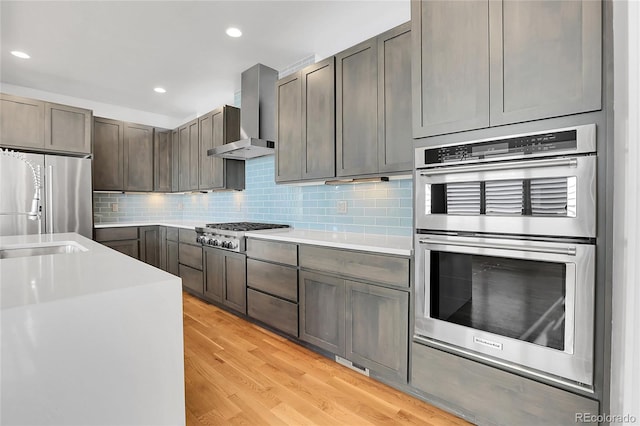 kitchen featuring decorative backsplash, stainless steel appliances, wall chimney range hood, and light hardwood / wood-style floors