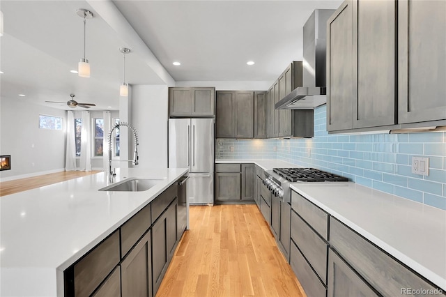 kitchen with sink, wall chimney exhaust hood, ceiling fan, light wood-type flooring, and appliances with stainless steel finishes